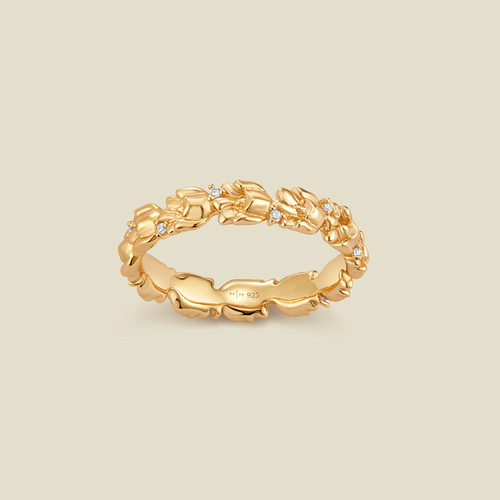 A gold floral-etched ring band with textured lily of the valley flowers and small clear gemstones, shown on a plain warm beige background. 