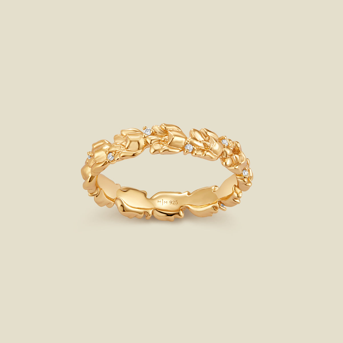 A gold floral-etched ring band with textured lily of the valley flowers and small clear gemstones, shown on a plain warm beige background. 