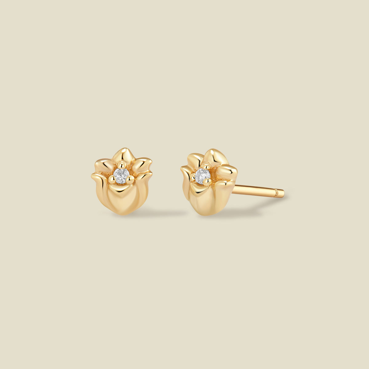 A pair of gold lily of the valley flower stud earrings, each featuring a small round clear gemstone at the center, displayed on a solid light beige background.