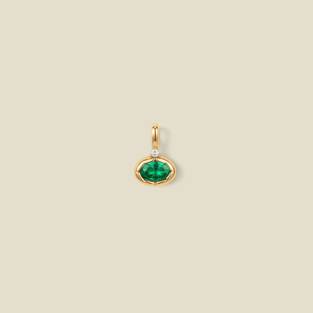 A gold oval pendant charm with a green bezel-set oval gemstone and a clear accent stone on top, set against a solid beige background.
