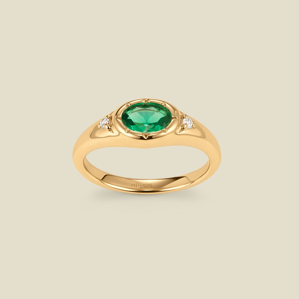 A gold signet ring with a green center oval bezel-set gemstone, and two small clear accent stones on either side, set against a solid beige background.