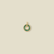 A gold circular pendant featuring dark green baguette-cut stones around a hollow center, displayed against a solid light beige background.