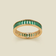 A gold eternity ring featuring a continuous channel setting of baguette-cut dark green gemstones, shown on a solid light beige background.