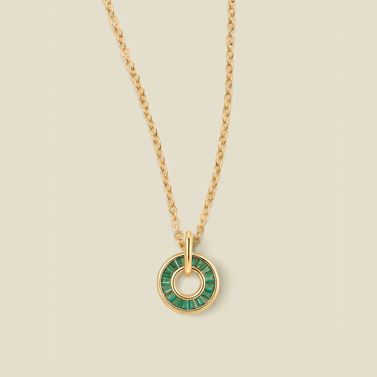 A gold chain necklace with a circular pendant featuring dark green baguette-cut stones around a hollow center, displayed against a solid light beige background.