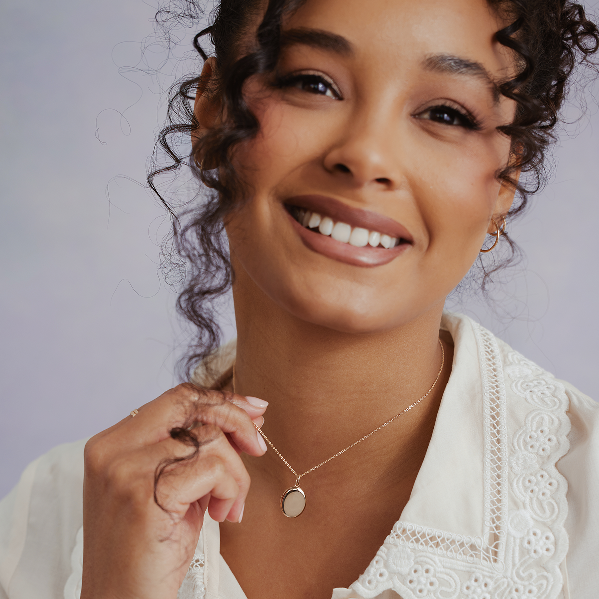 A close-up lifestyle shot of a smiling woman with curly hair wearing a delicate gold oval locket pendant necklace and a white eyelet blouse against a soft lavender-gray background.