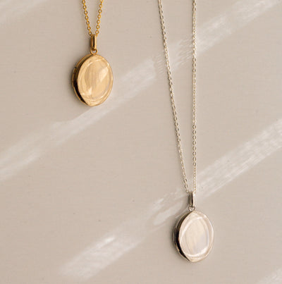 Two oval locket pendant necklaces displayed on a light beige surface with soft diagonal shadows. One necklace is gold and the other is silver.