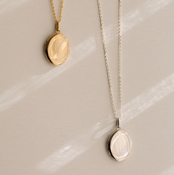 Two oval locket pendant necklaces displayed on a light beige surface with soft diagonal shadows. One necklace is gold and the other is silver.