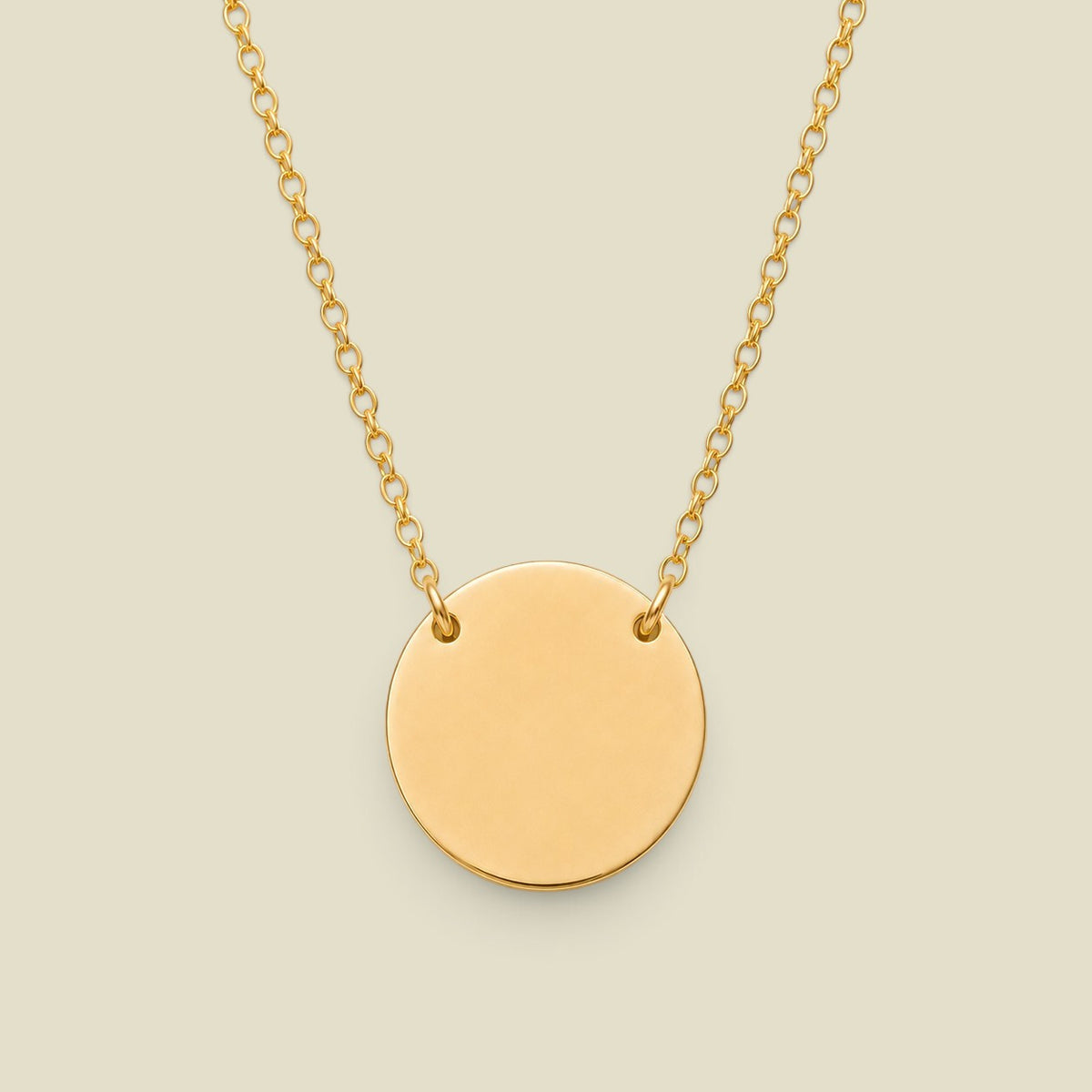 A minimalist gold disc pendant necklace attached to a delicate gold chain at two spots on the upper half of the disc, centered against a solid light beige background.