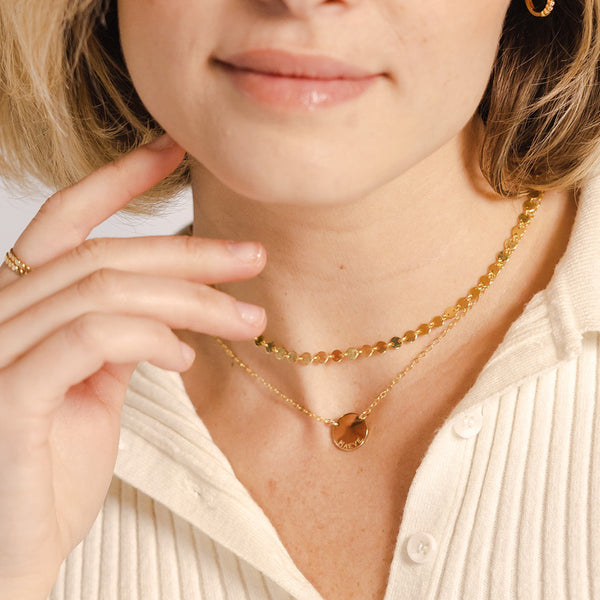 A close-up lifestyle shot of a model wearing two delicate gold layered necklaces, one with a small circular engraved pendant, paired with a cream ribbed knit top.