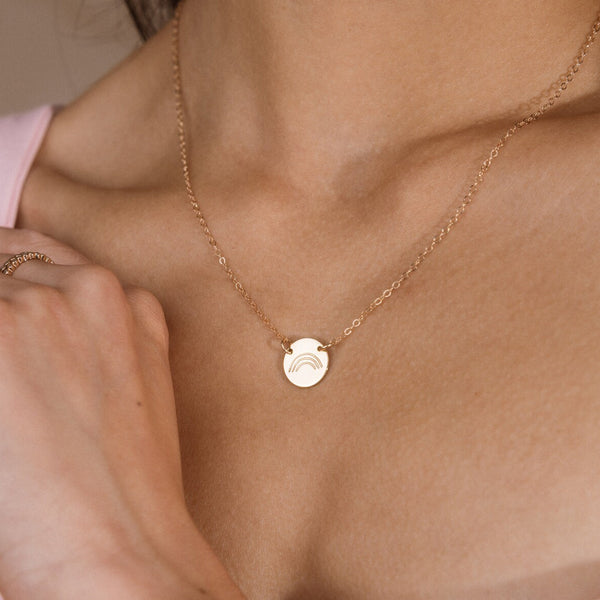A delicate gold chain necklace with a small round disc pendant featuring a subtle rainbow design, worn on a model's neck.