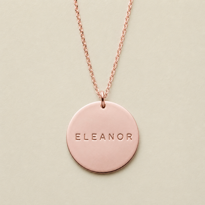 A rose gold round pendant necklace engraved with the name 'ELEANOR', displayed on a light beige background.