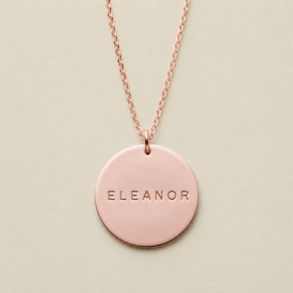 A rose gold round pendant necklace engraved with the name 'ELEANOR', displayed on a light beige background.