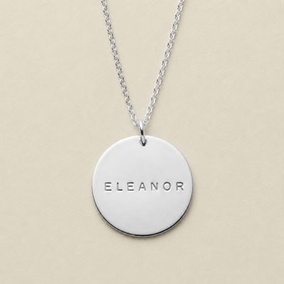 A silver round disc pendant necklace engraved with the name 'ELEANOR', suspended from a delicate silver chain against a solid beige background.