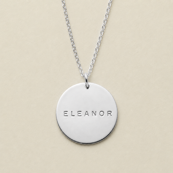 A silver round disc pendant necklace engraved with the name 'ELEANOR', suspended from a delicate silver chain against a solid beige background.