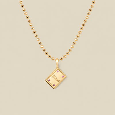 A gold ball chain necklace with a rectangular book charm featuring the word 'SMUT'  in gold lettering and four small pink gemstone accents, displayed on a solid beige background.
