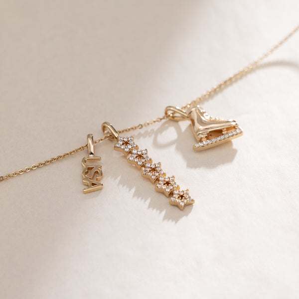 A gold dainty chain necklace featuring a 'USA' charm, a vertical stacking star charm with clear stones, and a figure skate charm.