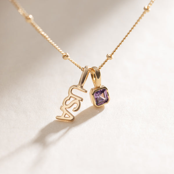 A gold dainty chain necklace with beads along it, holding a gold 'USA' charm and a square gold charm with a purple gemstone.