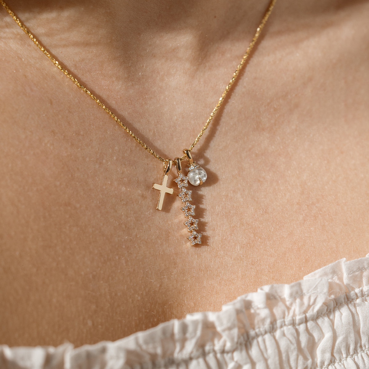 A close up lifestyle shot of a person's neck wearing a dainty gold chain necklace with a gold cross charm, a vertical stacked star charm with clear stones, and a ladybug charm with a clear stone.