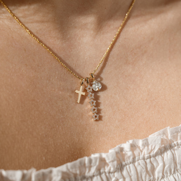 A close up lifestyle shot of a person's neck wearing a dainty gold chain necklace with a gold cross charm, a vertical stacked star charm with clear stones, and a ladybug charm with a clear stone.