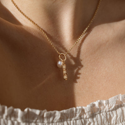 A close up lifestyle shot of a gold charm necklace with a round mechanism holding a round pearl charm and a gold snowboard charm.