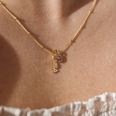 A close up lifestyle shot of a person's neck wearing a gold dainty chain necklace with beads along it, holding a gold 'USA' charm and a square gold charm with a purple gemstone.