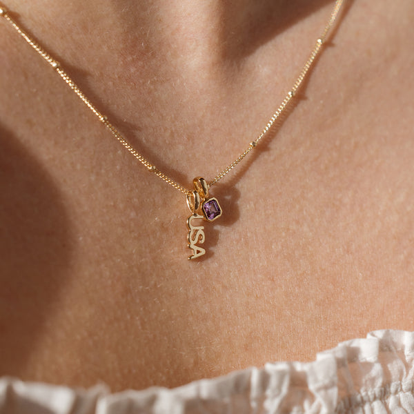 A close up lifestyle shot of a person's neck wearing a gold dainty chain necklace with beads along it, holding a gold 'USA' charm and a square gold charm with a purple gemstone.
