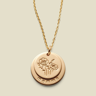 A gold disc pendant necklace featuring two overlapping discs on one jump ring, one with an engraved floral design, and the other larger disc underneath with the word 'momma' along the  bottom, displayed against a solid light beige background.