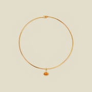 A thin gold wire choker necklace with a small oval amber gemstone pendant, displayed on a solid light beige background.