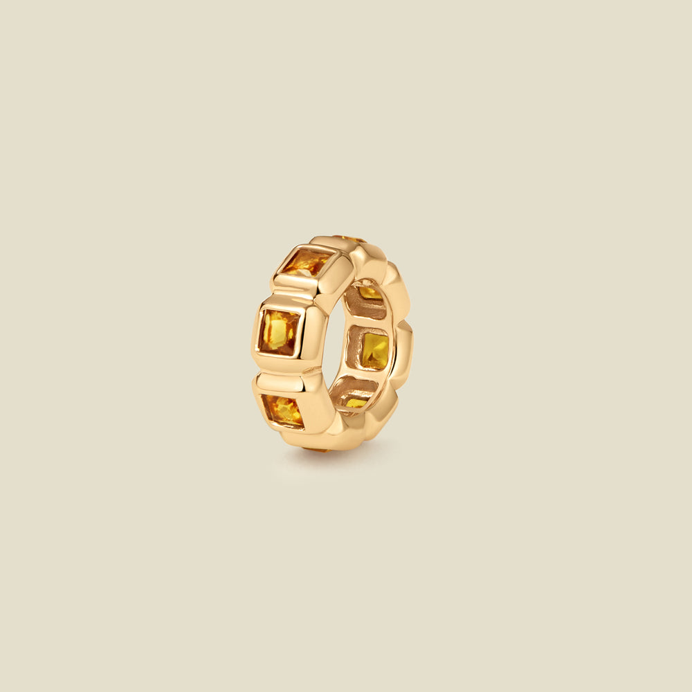 A gold ring charm with multiple bezel set square-cut amber gemstones set side by side to create the band, shown on a solid beige background.