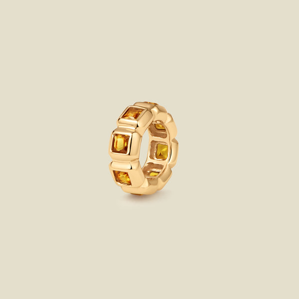 A gold ring charm with multiple bezel set square-cut amber gemstones set side by side to create the band, shown on a solid beige background.