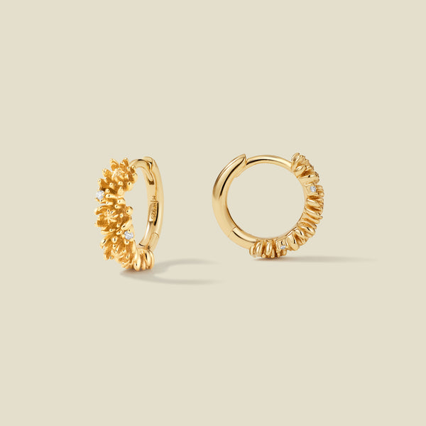A pair of gold hoop earrings adorned with chrysanthemum flowers stacked along the hoop, with scattered small clear accent stones, set against a solid beige background.