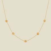 A delicate gold chain necklace featuring five small, evenly spaced textured chrysanthemum flower motifs set with tiny clear stones, displayed on a solid light beige background.