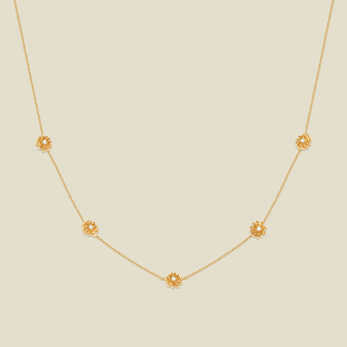 A delicate gold chain necklace featuring five small, evenly spaced textured chrysanthemum flower motifs set with tiny clear stones, displayed on a solid light beige background.