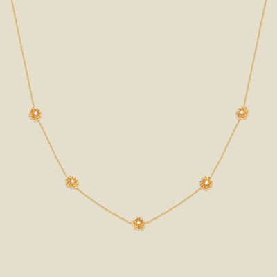 A delicate gold chain necklace featuring five small, evenly spaced textured chrysanthemum flower motifs set with tiny clear stones, displayed on a solid light beige background.