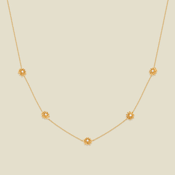 A delicate gold chain necklace featuring five small, evenly spaced textured chrysanthemum flower motifs set with tiny clear stones, displayed on a solid light beige background.