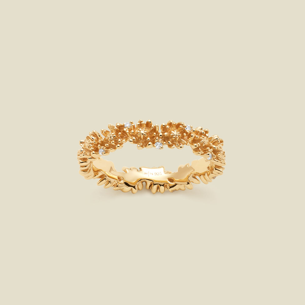 A gold floral-etched ring band with textured chrysanthemum flowers and small clear gemstones, shown on a plain warm beige background. 