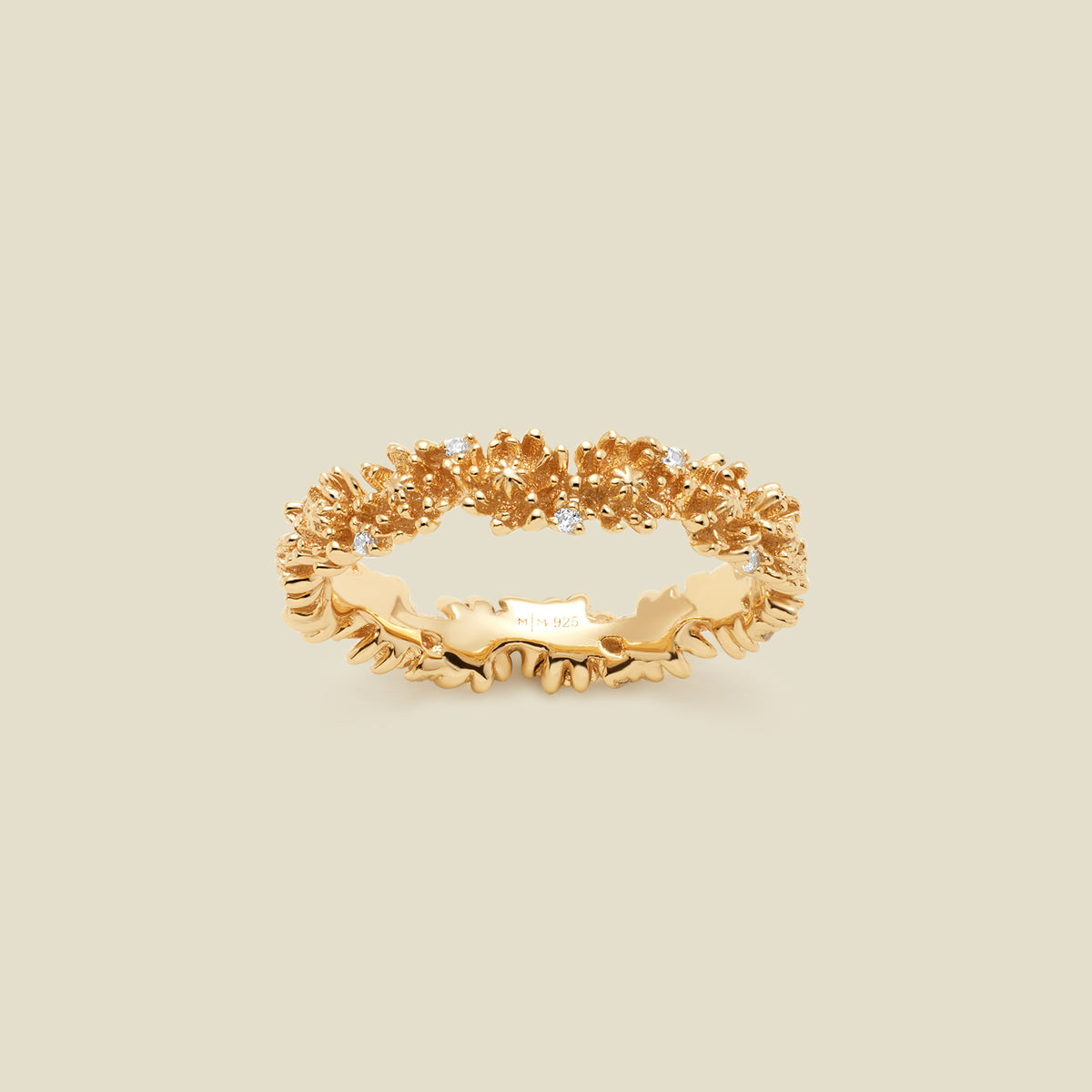 A gold floral-etched ring band with textured chrysanthemum flowers and small clear gemstones, shown on a plain warm beige background. 