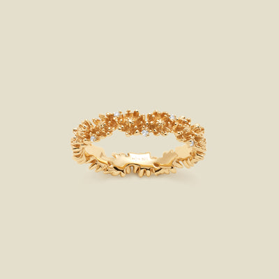 A gold floral-etched ring band with textured chrysanthemum flowers and small clear gemstones, shown on a plain warm beige background. 