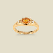 A gold signet ring with an amber center oval bezel-set gemstone, and two small clear accent stones on either side, set against a solid beige background.