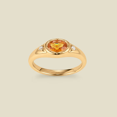 A gold signet ring with an amber center oval bezel-set gemstone, and two small clear accent stones on either side, set against a solid beige background.