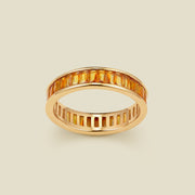 A gold eternity ring featuring a continuous channel setting of baguette-cut amber gemstones, shown on a solid light beige background.