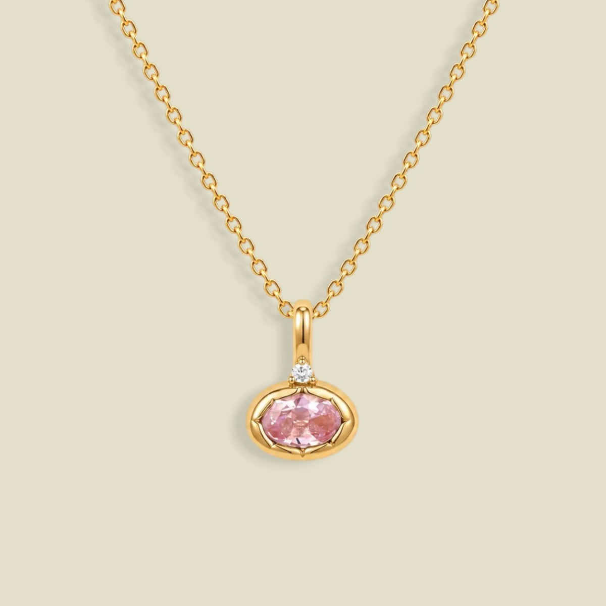 A delicate gold chain necklace with a small oval pendant featuring a pink gemstone and a tiny clear accent stone above it, set against a solid light beige background.