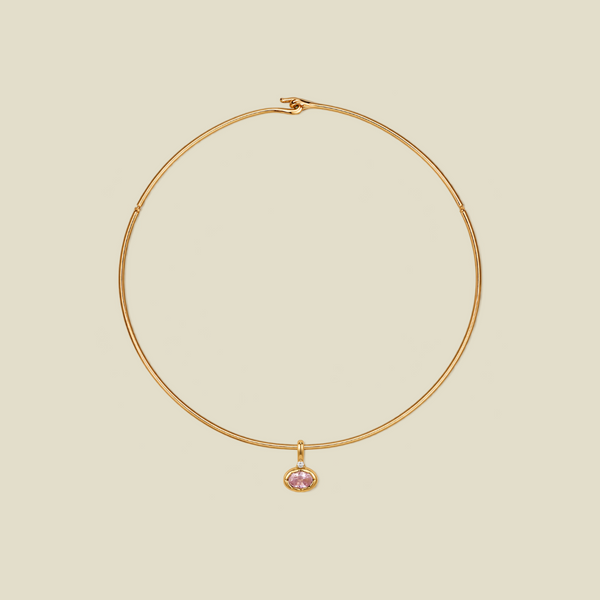 A delicate gold wire hoop necklace with a small oval-shaped pink gemstone pendant, displayed on a solid light beige background.