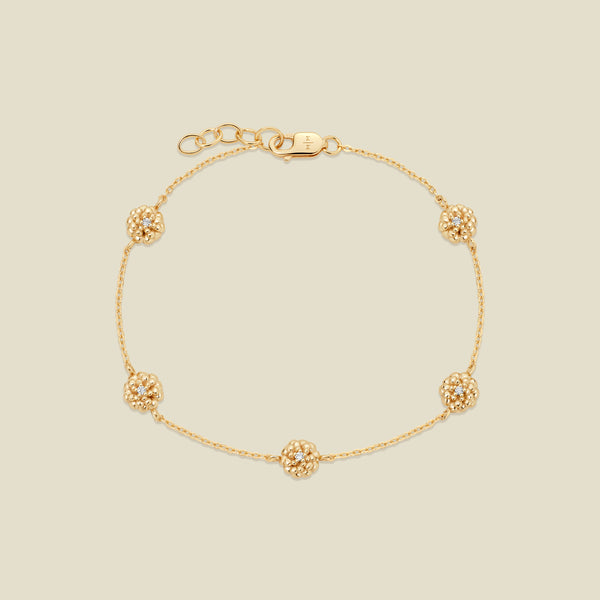 A delicate gold chain bracelet featuring five ornate, textured gold marigold charms with small clear stones, arranged in a circle on a light beige background.
