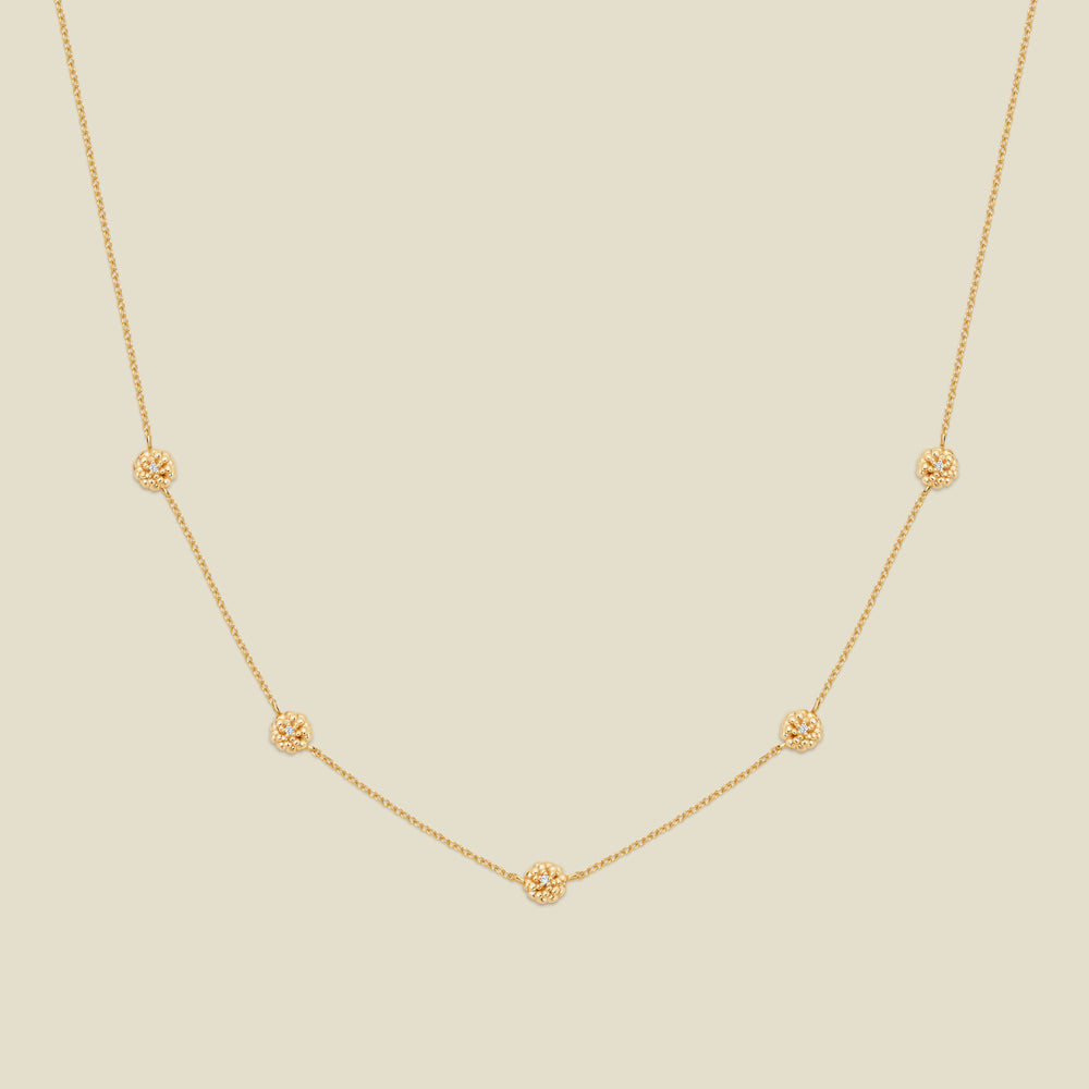 A delicate gold chain necklace featuring five small, evenly spaced textured marigold flower motifs set with tiny clear stones, displayed on a solid light beige background.