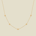 A delicate gold chain necklace featuring five small, evenly spaced textured marigold flower motifs set with tiny clear stones, displayed on a solid light beige background.
