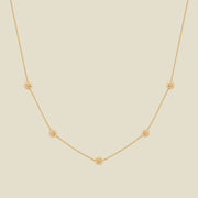 A delicate gold chain necklace featuring five small, evenly spaced textured marigold flower motifs set with tiny clear stones, displayed on a solid light beige background.