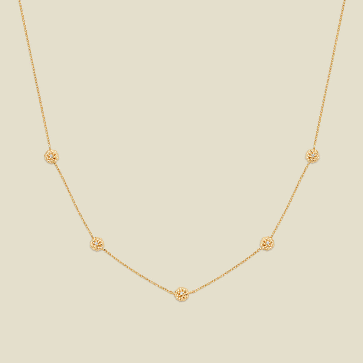 A delicate gold chain necklace featuring five small, evenly spaced textured marigold flower motifs set with tiny clear stones, displayed on a solid light beige background.