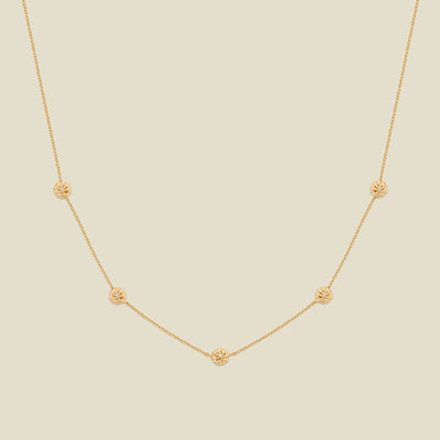 A delicate gold chain necklace featuring five small, evenly spaced textured marigold flower motifs set with tiny clear stones, displayed on a solid light beige background.
