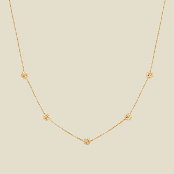 A delicate gold chain necklace featuring five small, evenly spaced textured marigold flower motifs set with tiny clear stones, displayed on a solid light beige background.